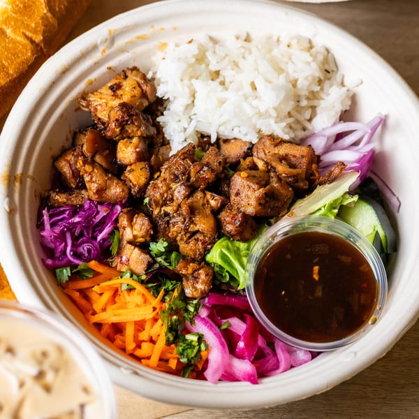 A takeout bowl with rice, grilled chicken, shredded carrots, red cabbage, pickled onions, and sauce.