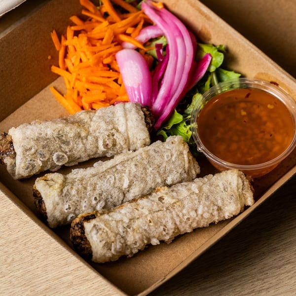 Three crispy spring rolls with shredded carrots, pickled onions, lettuce, and dipping sauce in a takeout box.
