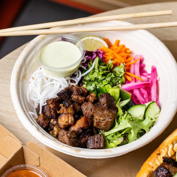 A bowl of beef, noodles, and vegetables with a side of sauce, chopsticks, and iced tea.