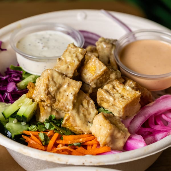 A close-up of a vibrant salad bowl with tofu, pickled onions, carrots, cucumbers, and two sauces.